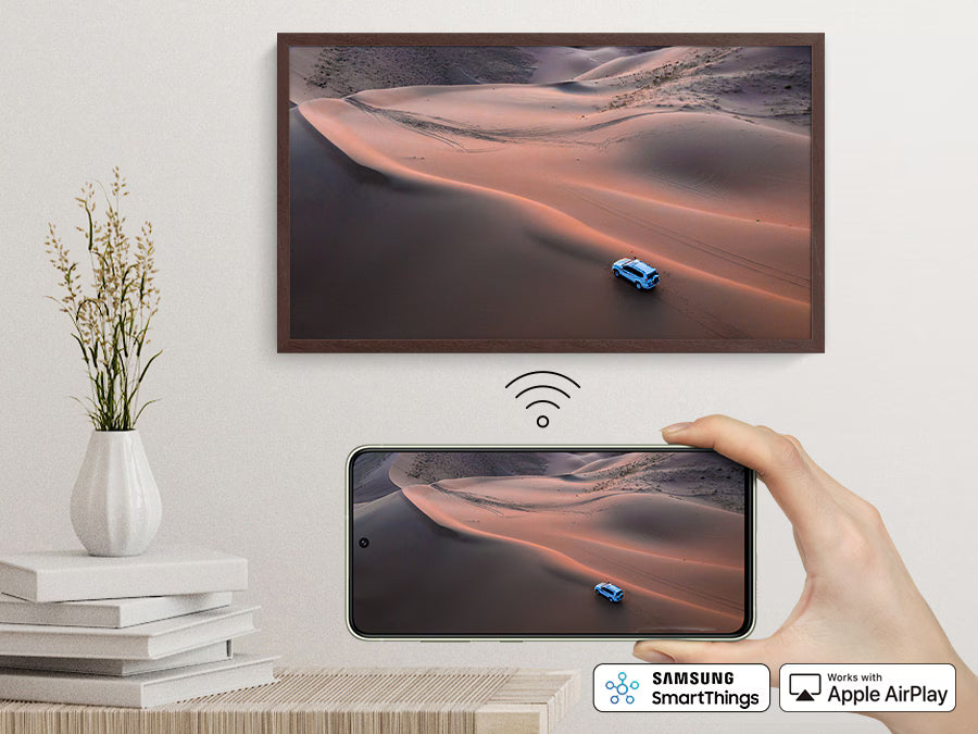 Smartphone displaying a desert scene with a car, connected to a larger screen above, on a light-colored wall.